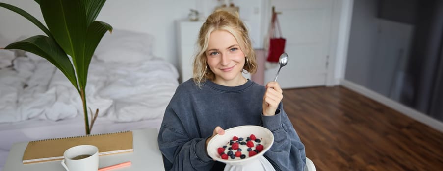 Portrait of cute smiling blond woman eats breakfast in her bedroom, looking at camera, holding bowl with dessert and spoon.
