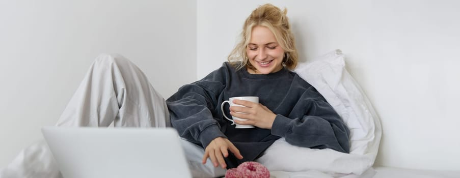 Portrait of happy blond girl, female model lying in bed in cosy clothes, eating and watching movie on laptop, smiling while looking at screen.