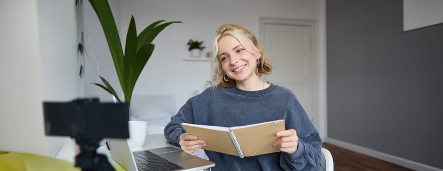 Portrait of smiling blond woman, sitting in bedroom, using laptop and digital camera, recording video for lifestyle blog, reading, using her notebook.