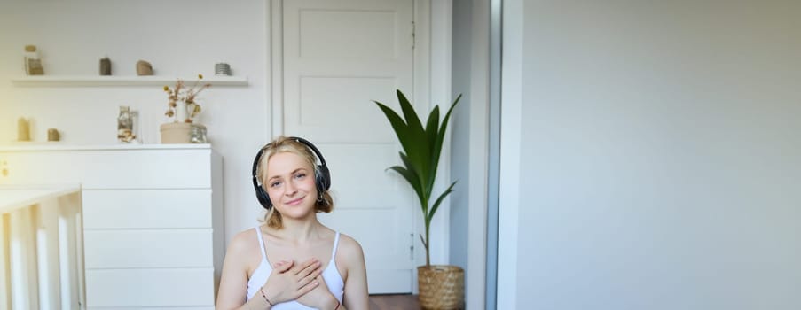 Vertical portrait of beautiful young woman in headphones, sits on yoga mat, meditating, holding hands on her chest and smiling at camera.