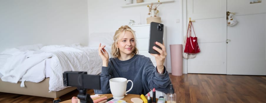 Portrait of young beauty blogger, recording video on digital camera, doing online live stream while applying makeup, talking to her followers, sitting on bedroom floor.