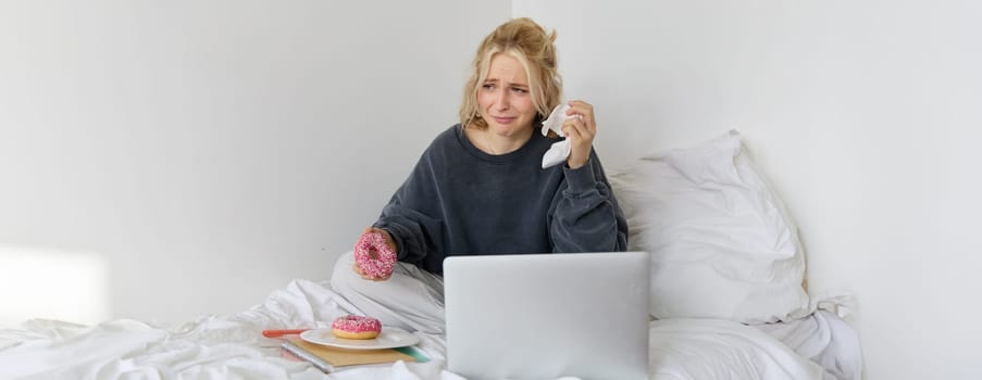 Portrait of woman sitting on a bed with laptop, eating doughnut and crying from sad movie scene.