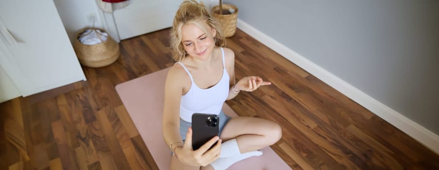 Portrait of woman having live stream training session, using her smartphone to create workout content, blogging during exercises or doing yoga, sitting on rubber mat at home.