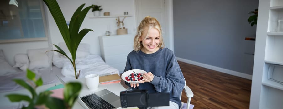 Portrait of smiling girl vlogger, blogger holding bowl with dessert, showing it on camera to audience, recording vlog in room.