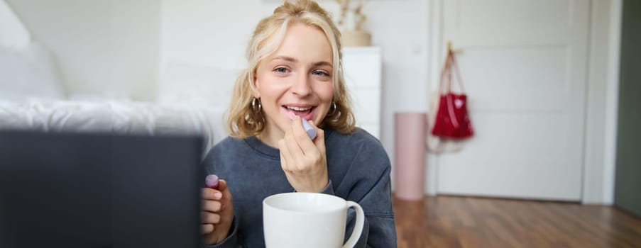 Lifestyle, beauty blogger, woman recording video of her putting on makeup, talking to camera, making online tutorial, showing her lip gloss or lipstick to followers, sitting on floor with cup of tea.