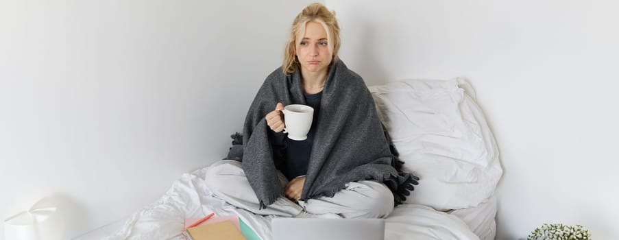 Portrait of woman catching a flu, sneezing, feeling sick, sitting on bed with laptop and working on remote while caught a cold.