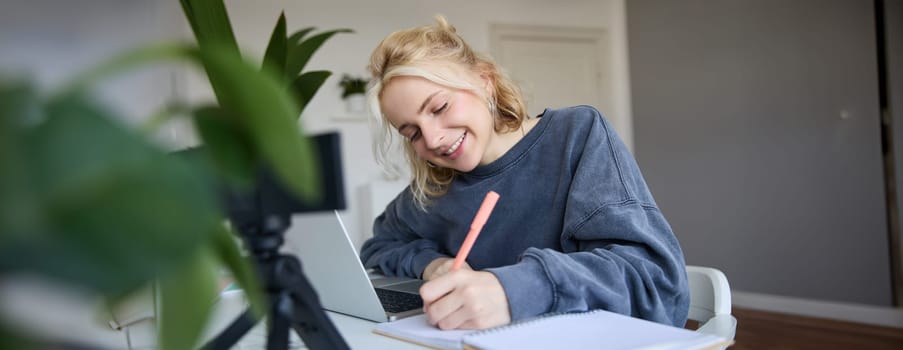 Portrait of young woman, lifestyle blogger, recording video of herself, making notes, writing in journal, sitting in front of laptop in a room and studying.