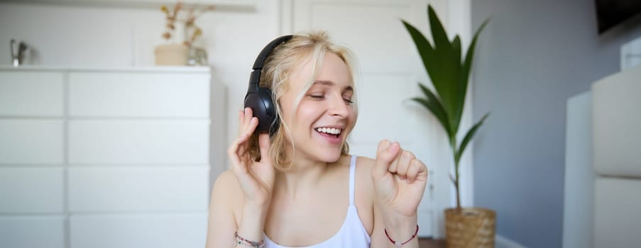 Portrait of happy woman, listens to music in wireless headphones, holding invisible microphone, singing into mic, smiling, enjoying good sound quality.