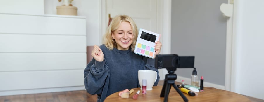 Excited smiling blond woman recording video review on a new eyeshadow palette, lifestyle blogger showing product to followers, sits on floor with enthusiastic face.