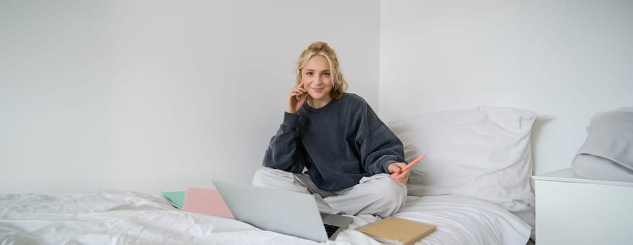 Portrait of young smiling woman studying in her bed, working from home in bedroom, sitting with laptop and notebooks on lotus pose, looking happy and relaxed.