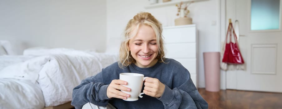 Portrait of young blond woman in casual clothes, sits on bedroom floor with cup of tea, drinking and looking at camera.