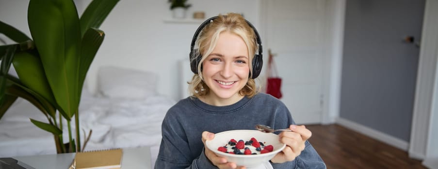 Portrait of smiling cute woman in headphones, eating her breakfast and watching video on laptop, looking at screen with happy face.