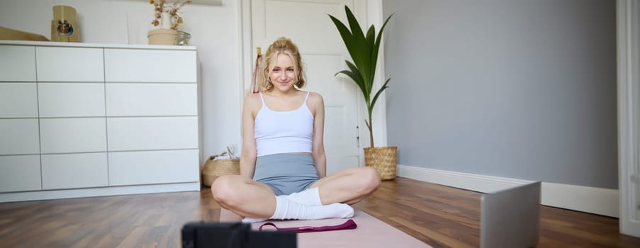 Portrait of young fitness instructor, vlogger showing exercises on camera, recording herself, sitting on mat with laptop, doing workout, explaining yoga movements to followers.