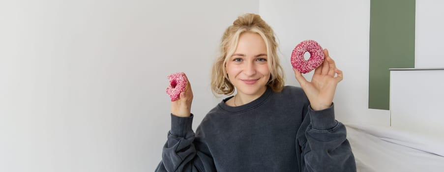 Close up portrait of happy, cute blond woman, holding doughnut, eating sweet, delicious comfort food, showing dessert at camera. Food concept