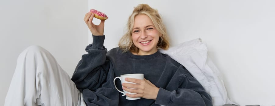 Close up portrait of beautiful smiling, blond woman, lying in bed, drinks tea and eats doughnut on a lazy weekend or day-off.
