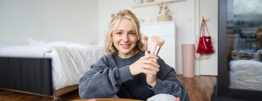 Portrait of young woman, content creator, making a video about makeup, showing brushes to audience, looking at camera, recording beauty tutorial, smiling happily.