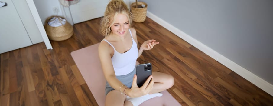 Portrait of young blogger, fitness instructor showing how to do exercises, sitting on yoga mat with smartphone, talking to followers on live stream.