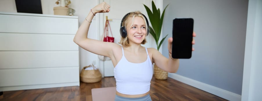 Portrait of beautiful and fit young woman, staying healthy working out at home, showing mobile app, blank screen, flexing biceps, recommending smartphone application for exercises.