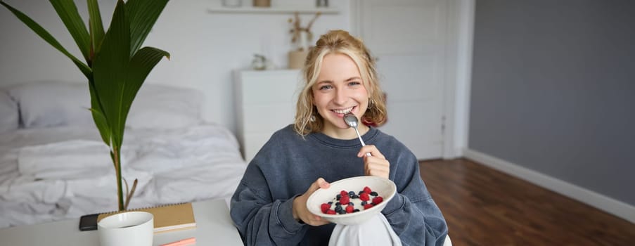 Portrait of beautiful young woman in a room, eating breakfast, holding bowl with dessert and a spoon, smiling at camera, recording lifestyle vlog.