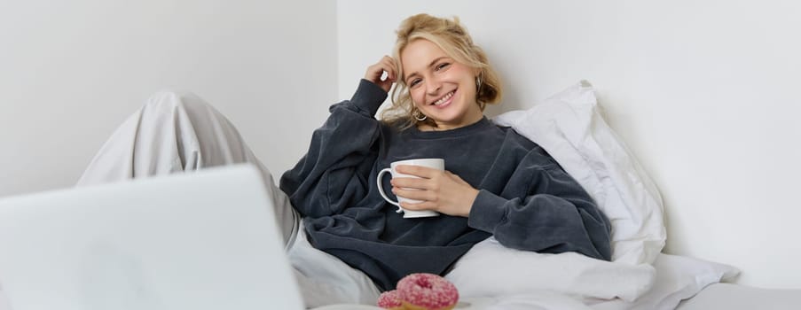 Portrait of happy blond girl, female model lying in bed in cosy clothes, eating and watching movie on laptop, smiling while looking at screen.