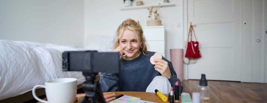 Image of young woman, vlogger records video on digital camera, shows beauty products, recommending makeup for audience on social media, sits on front in a room.