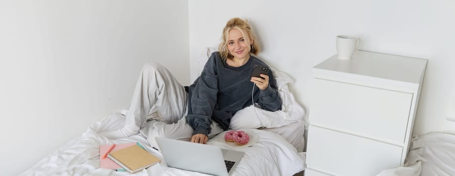 Portrait of young woman, student studying in her bed, relaxing while preparing homework, eating doughnut, using laptop in bedroom and drinking tea.