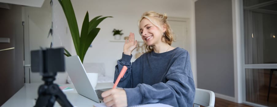 Portrait of young blond smiling woman, studying at home, remote education concept, connects to online course or lesson, writing in notebook.