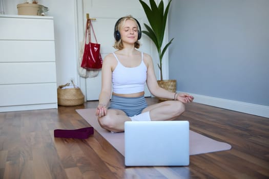 Portrait of concentrated woman in headphones, meditating, listening to calm music and practice yoga, follow instructions on laptop, sitting on rubber mat.