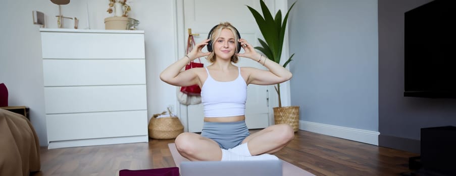 Portrait of young woman at home, connects to online gym training session, following fitness instructions on laptop, wearing wireless headphones during workout.