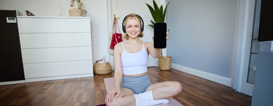Portrait of beautiful young fitness woman, wearing headphones, showing workout app on smartphone, demonstrating mobile phone screen and smiling, sitting on yoga mat.