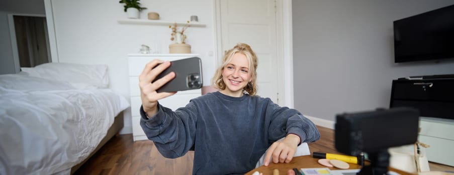 Image of young woman, vlogger taking selfie in her room, talking to her followers during online live stream, using smartphone app to chat with audience, smiling and looking happy.