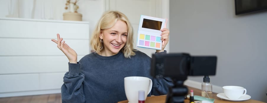 Portrait of beautiful lifestyle blogger, girl records a video on her camera for social media, shows palette of eyeshadows, does a makeup tutorial for her followers, sits in her room.