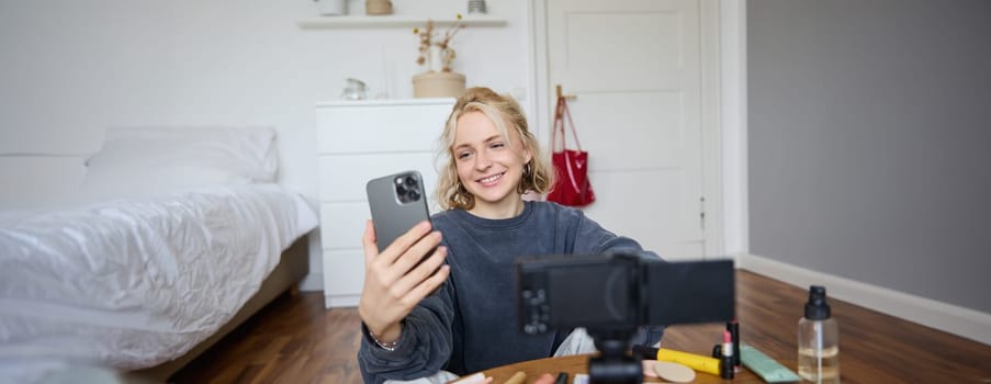 Portrait of young beautiful woman, lifestyle vlogger, sitting on bedroom floor, recording video on digital camera, using smartphone for online live stream, chatting with audience, reading messages.