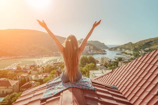 Woman sits on rooftop with outstretched arms, enjoys town view and sea mountains. Peaceful rooftop relaxation. Below her, there is a town with several boats visible in the water.