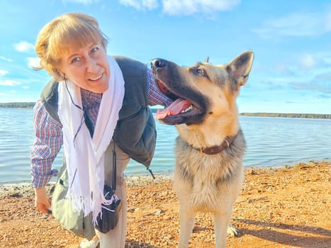 Adult girl with shepherd dog taking selfie near water of lake or river. Middle aged woman and big pet on nature. Friendship, love, fun, hugs