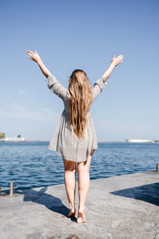 woman in a dress stands on a dock by the water, her arms raised in the air. Concept of freedom and joy, as the woman is celebrating or expressing her happiness