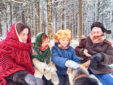 Joyful family ethnic dress with shawls and earflap hats and dog in winter forest in Russia carnival Maslenitsa. Tourists in Shrovetide in spring. Mother, father, son, daughter having fun in snow