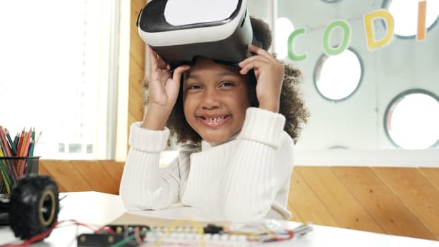 African girl taking off VR glass or head set while looking at camera. Student smiling at camera with colored pencil and laptop placed on table in STEM technology class. Online education. Erudition.