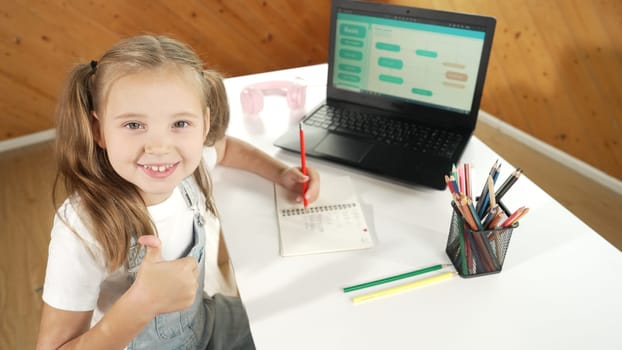 Little pretty caucasian girl doing classwork while showing a thumb up gesture. Young child writing plan in note book while looking at engineering programing code in STEM technology class.. Erudition.
