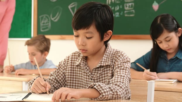 Asian attractive boy taking a note while student writing answer in answer sheet. Multicultural student doing classwork or test while caucasian teacher checking student homework at classroom. Pedagogy.