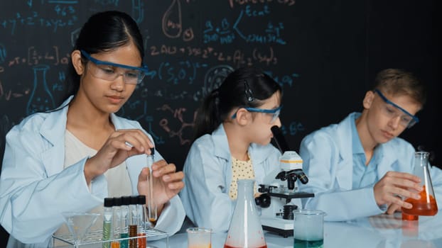 Cute girl looking under microscope while student doing experiment at blackboard with theory written. Young scientist inspect colored solution at table with experimental equipment placed. Edification.