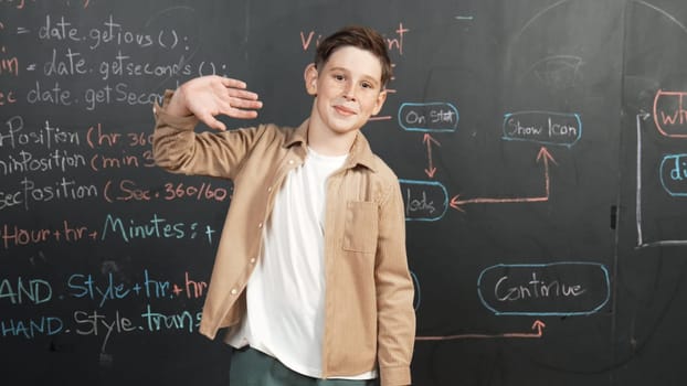 Panorama shot of happy caucasian boy smiling and waving hand at blackboard with engineering code or prompt written. Smart child looking at camera and greeting while study in STEM classroom. Erudition.