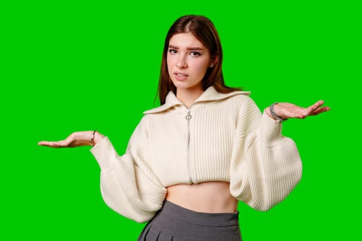 A young woman stands against a vibrant green backdrop, her expression one of uncertainty or confusion. With her arms outstretched and palms facing upwards, she embodies a visual shrug, symbolizing indecision or lack of knowledge about a particular situation.