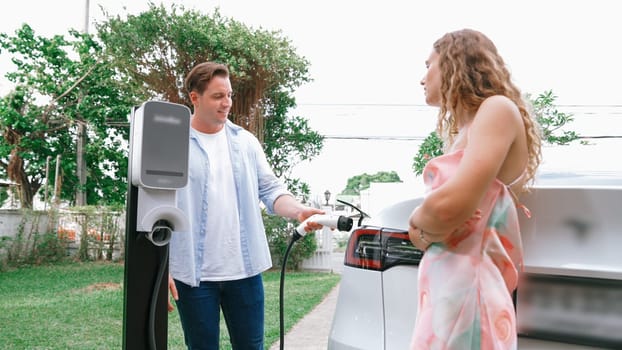 Happy and lovely couple with eco-friendly conscious recharging electric vehicle from EV charging station. EV car technology utilized as alternative transportation for future sustainability. Synchronos