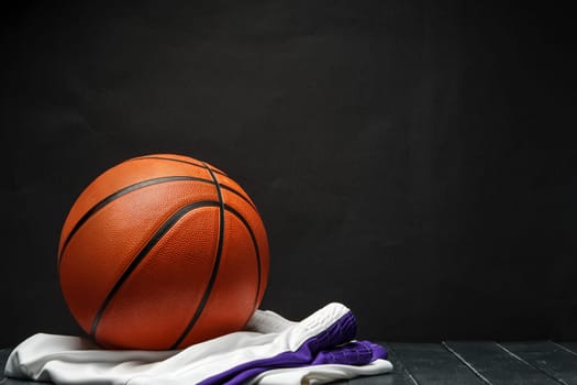 A freshly polished orange basketball sits on a dark wooden surface, contrasting with the deep black background. A white uniform with purple trim is casually draped next to the ball, suggesting a pause in play or the aftermath of a game.
