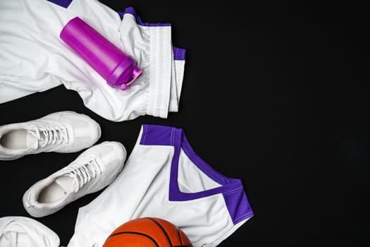 A collection of basketball essentials is neatly arranged against a stark, dark background, including a vibrant orange basketball, white sneakers with purple detailing, a white jersey with purple accents, and a purple water bottle, suggesting readiness for an athletic training session or a game.