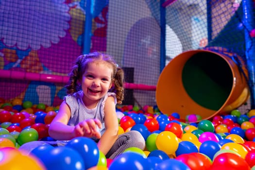 A young girl with a beaming smile sits amongst a sea of vibrant multicolored balls in an indoor play area. Her casual attire suggests a day of fun and activity as she reaches out, enjoying the playful environment around her. The background features soft play structures and a tunnel slide, highlighting the energetic atmosphere of the childrens playground.