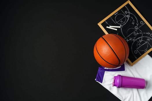 A basketball, along with a strategic play diagrammed on a chalkboard, rests near a folded jersey and a sports bottle. The equipment is arranged on a dark surface, suggesting preparation for a game or a coaching session in progress.