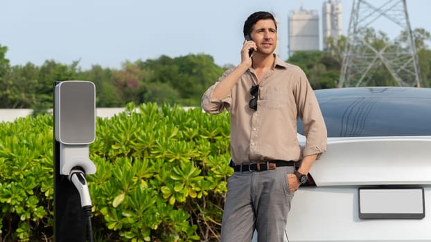 Man talking on the phone while recharge EV car battery at charging station connected to power grid tower electrical as electrical industry for eco friendly car utilization. Panorama Expedient