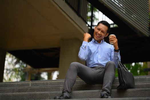 Happy young businessman celebrating success, expressing positivity while sitting outside office building.